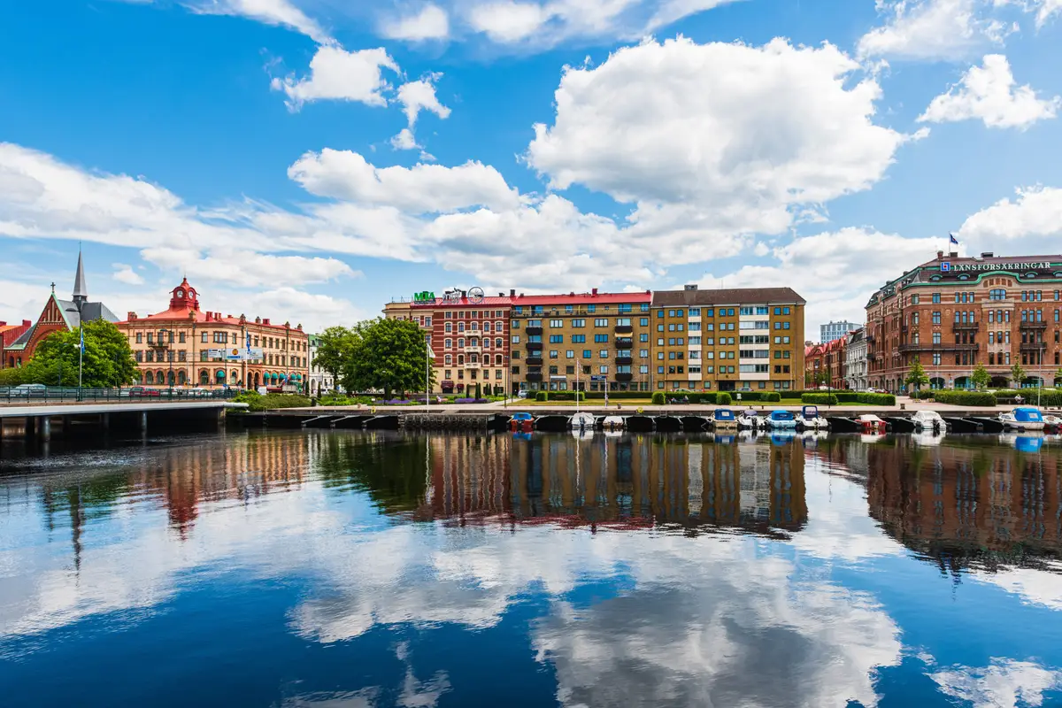Colorful buildings of Halmstad city along a calm river in Sweden, with reflections visible in the water and a clear blue sky with clouds above.