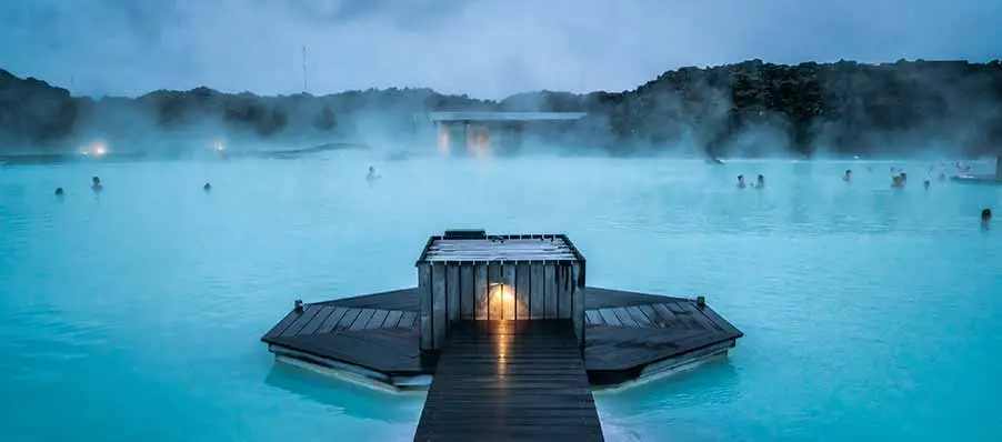Tourists enjoying a serene evening at the geothermal Blue Lagoon spa in Iceland, with steam rising over tranquil blue waters and a wooden walkway leading to a cozy lit hut.
