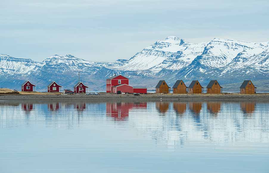 Picturesque East Iceland cottages with red roofs reflected in calm water, scenic mountain backdrop in early spring.