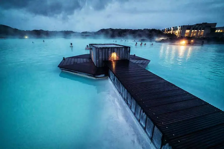 Visitors relaxing in the steamy milky-blue waters of the geothermal spa, Blue Lagoon, during twilight in Reykjavik, Iceland, highlighting the spa's serene ambiance and unique landscape.