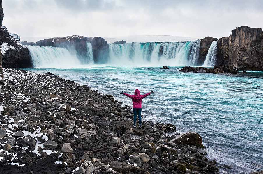 Tourist in pink jacket admiring the majestic Godafoss Waterfall in Iceland, surrounded by icy rocks and turbulent blue waters.