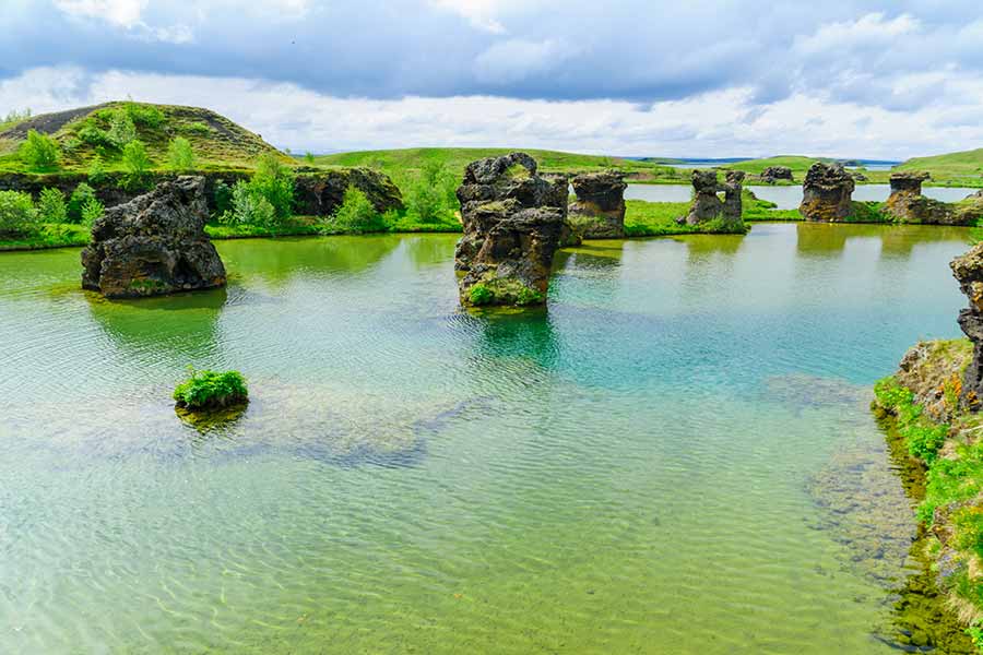 Scenic view of volcanic rock formations at Lake Mývatn, Iceland, showcasing lush greenery and clear blue waters, ideal for nature and travel enthusiasts.