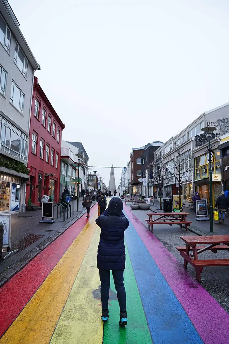 Tourist walking on colorful rainbow road at Laugavegur Street, Reykjavik, Iceland, with shops and Hallgrimskirkja church in the background.