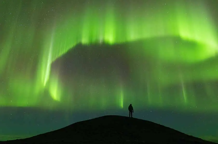 A lone observer stands on a hill under the dazzling green aurora borealis in the night sky of Iceland.