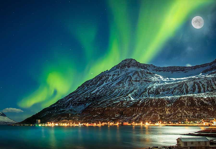 Vibrant Northern Lights over snowy Seydisfjordur village in Iceland under a full moon