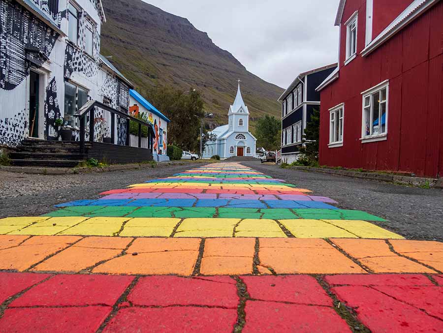 Colorful rainbow-painted road leading to a quaint church in Seydisfjordur Village, Iceland, surrounded by traditional Nordic buildings and a mountainous backdrop.