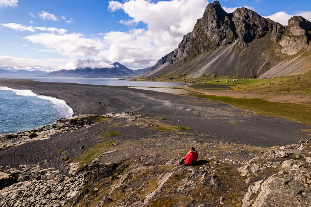 Couple in red jackets enjoying the panoramic views of Hvalnes Nature Reserve with its distinctive black beach and dramatic mountain backdrop in Iceland