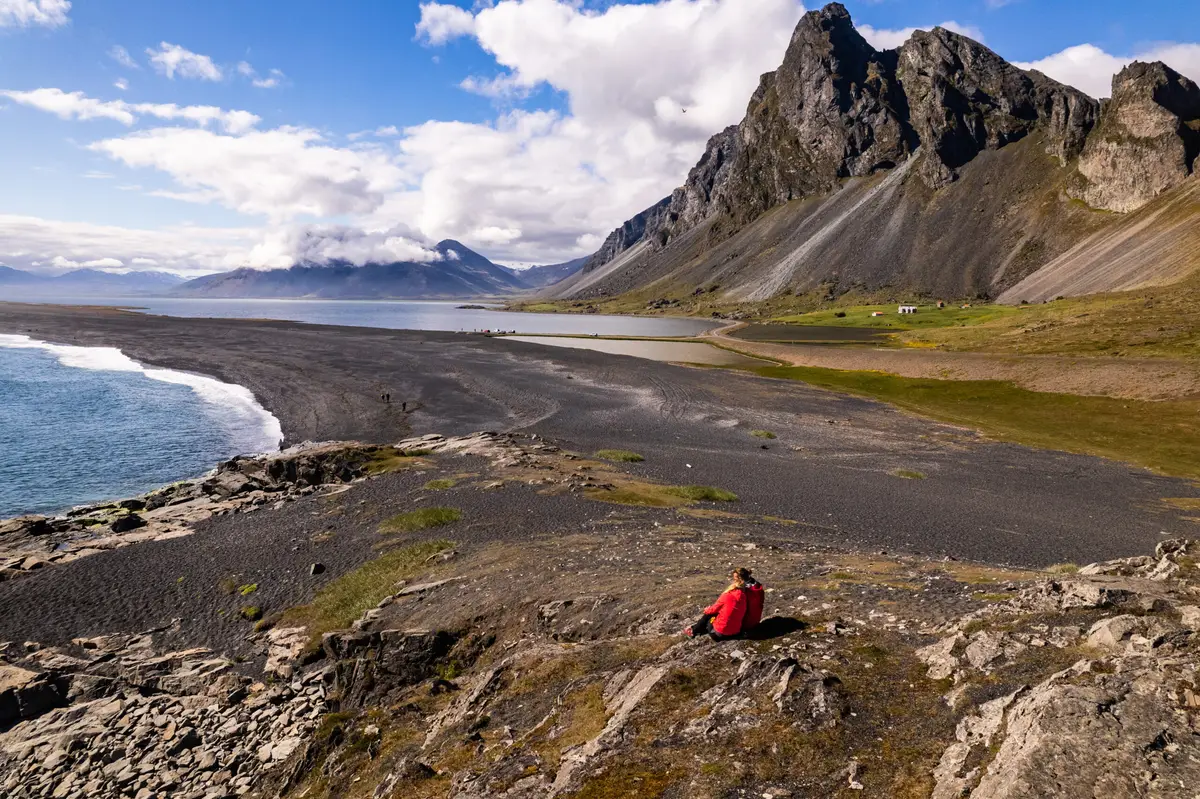 Couple in red jackets enjoying the panoramic views of Hvalnes Nature Reserve with its distinctive black beach and dramatic mountain backdrop in Iceland