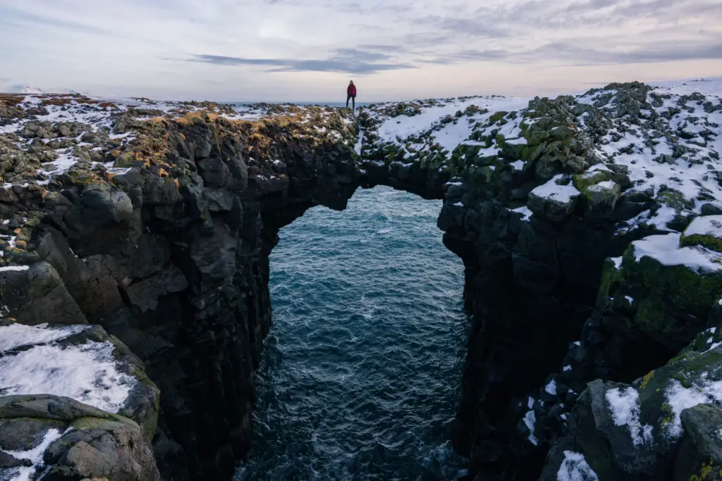 Person standing on a natural arch over the icy sea in Arnarstapi, Snaefellsnes Peninsula, Iceland, highlighting dramatic winter coastline scenery.