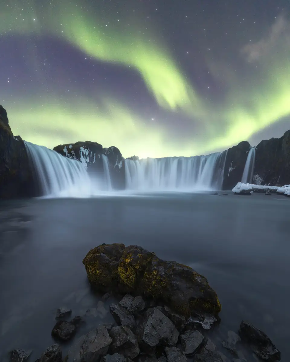 Majestic Aurora Borealis over a serene waterfall in Iceland, showcasing vibrant green northern lights in a starry sky with cascading water and rugged rocks in the foreground.