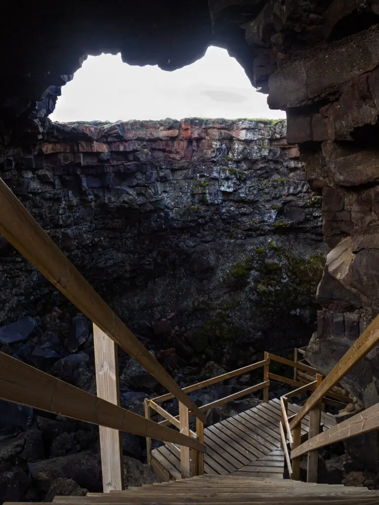 Wooden stairs descending into the Víðgelmir lava tube cave in Borgarfjörður, Iceland, showcasing rugged volcanic rock formations and natural cave openings.